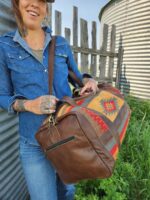 A person wearing a denim shirt and blue jeans carries a large, colorful duffel bag with patterns and leather accents. They stand beside a corrugated metal wall and a wooden fence, with green grass and a round metal structure in the background.