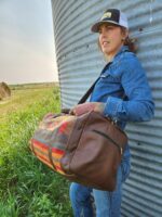 A person wearing a denim outfit and a baseball cap stands next to a large metal cylinder, holding a brown and multicolored patterned duffle bag. They are outdoors in a grassy field with hay bales visible in the background.