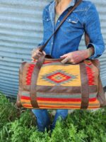 A person dressed in a denim shirt and blue jeans holds a large, rectangular bag with a tribal pattern and leather straps. The background features a corrugated metal wall and green grass. The person's arms are covered in colorful tattoos.