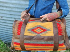 A person dressed in a denim shirt and blue jeans holds a large, rectangular bag with a tribal pattern and leather straps. The background features a corrugated metal wall and green grass. The person's arms are covered in colorful tattoos.