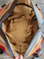 A brown leather handbag with a multicolored striped design on the outside is open to reveal a spacious, tan interior. The inside features a zippered pocket on one side and two open pockets on the other. The bag is held open by a hand at the bottom.