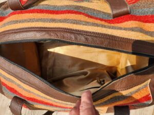 A close-up view of an open handbag featuring brown leather handles and vibrant striped fabric in orange, yellow, and red hues. The interior of the bag is empty, showcasing a beige lining and a small inner pocket. The bag is being held open by a hand.