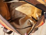 A top-down view of an open, mostly empty tote bag with a brown fabric interior and a textured brown, red, and orange exterior. The bag has several zippered and unzippered pockets, and one hand is holding the bag open. The bag is on a wooden surface.