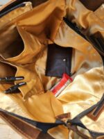 The image shows the interior of a tan bag. Inside are various compartments, some of which are filled with items. Visible items include three pens, a brown wallet, and a red snack bar. The lining of the bag is golden, with an organized layout.