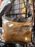 A brown and white cowhide leather handbag with shoulder straps is displayed hanging on a wire rack. The bag has an uneven color pattern with patches of white and shades of brown. The background shows other leather goods and a bright light in the corner.