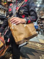 A person wearing a black shirt with red floral embroidery is carrying a brown and white cowhide bag with a shoulder strap. They are standing beside displayed rugs and leather goods in what appears to be a market or shop.