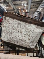A handbag with a brown leather strap and top border is displayed. The body of the bag features a white and brown speckled cowhide pattern. The bag is hanging on a metal rack in a store, with other items visible in the background.
