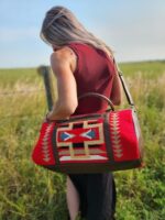 A woman with long blonde hair stands in an open field with tall grass, facing away from the camera. She wears a sleeveless maroon top and carries an intricately patterned red and brown bag with geometric designs over her shoulder. A wooden fence is visible in the background.