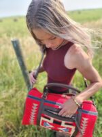 A woman with blonde hair and pink highlights stands in a field with a blurry background. She is looking down and carrying a large red-patterned duffel bag over her shoulder with both hands. She wears a maroon sleeveless top and a bracelet on her left wrist.