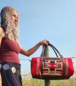 A woman with long, wavy hair stands outdoors against a clear blue sky, dressed in a maroon sleeveless top and a black skirt adorned with silver coins. She rests her arm on a wooden post, showcasing a red and black patterned handbag.