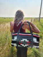 A person with long hair stands in a grassy field, facing away from the camera. They are wearing a burgundy top and holding a black, white, and red patterned duffel bag over their shoulder. A fence runs alongside them in the background.