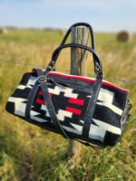 A duffel bag with black handles and strap, featuring a bold geometric pattern in black, white, and red, hangs on a wooden fence post. The background is a grassy field with hay bales and clear blue sky.