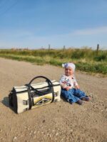 A baby wearing a white hat with a bow and a patterned sweater, sitting on a dirt road. Next to the baby is a black and white patterned bag. The background features a clear blue sky, green grass, and a wooden fence.