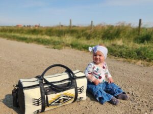 A baby wearing a white hat with a bow and a patterned sweater, sitting on a dirt road. Next to the baby is a black and white patterned bag. The background features a clear blue sky, green grass, and a wooden fence.