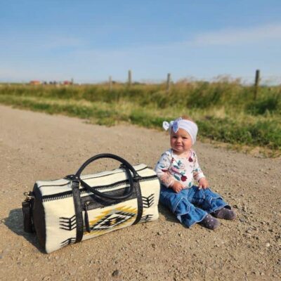 A baby wearing a white hat with a bow and a patterned sweater, sitting on a dirt road. Next to the baby is a black and white patterned bag. The background features a clear blue sky, green grass, and a wooden fence.