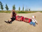 A woman with long hair and a baby sit on a dirt road next to two red tote bags with geometric patterns. The baby, wearing a headband and jeans, reaches toward one of the bags. Trees line the road in the background under a clear blue sky.