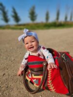 A smiling baby wearing a headband with a bow, sitting in a brown leather bag lined with a red, patterned blanket, on a dirt ground. The background features a clear blue sky and blurred, tall trees.