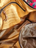 A close-up view of the inside of an open bag, showing a shiny, gold-colored fabric lining and a zippered pocket with a leather pull tab. The top edge of the bag has a plaid fabric accent visible in the top right corner.