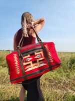 A person with long blonde hair, wearing a maroon top, carries a large, vibrant red bag with geometric patterns over their shoulder. They are standing in an open field, with a clear blue sky in the background.