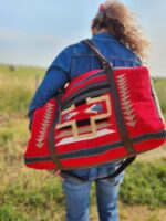 A person with long hair, wearing a denim jacket and blue jeans, is seen from behind carrying a large red, patterned bag with black and white geometric designs on their back. They are walking outdoors in a grassy field on a sunny day.