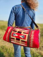 A person wearing a denim shirt faces away from the camera, standing in a grassy field. They carry a bright red duffel bag with a colorful southwestern pattern and brown leather straps slung over their shoulder.