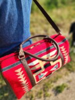 A person wearing blue jeans is carrying a vibrant red duffel bag with a Southwestern geometric pattern and brown leather straps. The background is blurred greenery.