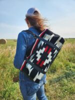 A person wearing a denim jacket and cap walks through a grassy field, carrying a patterned blanket with black, white, and red geometric designs strapped to their back. Hay bales and a clear blue sky are visible in the background.