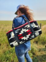 A person with long hair, wearing a blue denim jacket and jeans, faces away from the camera in an open field. They are carrying a bag with a Native American-inspired geometric pattern in black, white, red, and gray.