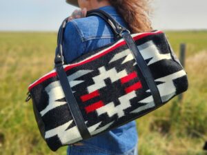 A person with long hair, wearing a blue denim jacket and jeans, faces away from the camera in an open field. They are carrying a bag with a Native American-inspired geometric pattern in black, white, red, and gray.