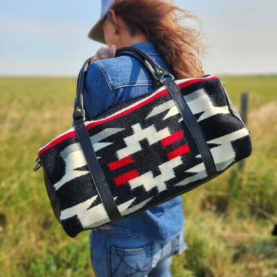A person with long hair, wearing a blue denim jacket and jeans, faces away from the camera in an open field. They are carrying a bag with a Native American-inspired geometric pattern in black, white, red, and gray.
