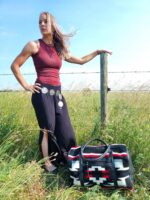 A woman with long hair stands in a field next to a wooden post and barbed wire fence, wearing a maroon sleeveless top, black skirt, and a decorative belt. A patterned travel bag rests on the grass. The sky is bright blue with some scattered clouds.