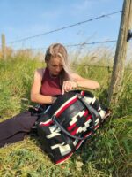 A woman with long hair sits in a grassy area by a barbed wire fence, wearing a red sleeveless top and dark pants. She is looking into a large black, white, and red patterned bag beside her. It's a bright and sunny day with blue skies.