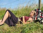 A woman with long hair is lying on the grass in a field, resting her head on a woven blanket. She is wearing a sleeveless top and a long skirt, with a bracelet on her wrist. She looks relaxed, surrounded by tall grass and a barbed wire fence in the background.