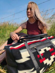 A woman with long hair sits in a grassy area near a barbed wire fence under a clear blue sky. She is wearing a burgundy tank top and is holding a large patterned duffel bag with black, white, and red colors on her lap. She appears to be deep in thought.