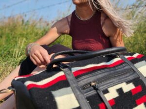 A woman with long hair sits in a grassy area near a barbed wire fence under a clear blue sky. She is wearing a burgundy tank top and is holding a large patterned duffel bag with black, white, and red colors on her lap. She appears to be deep in thought.