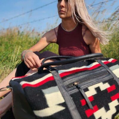 A woman with long hair sits in a grassy area near a barbed wire fence under a clear blue sky. She is wearing a burgundy tank top and is holding a large patterned duffel bag with black, white, and red colors on her lap. She appears to be deep in thought.
