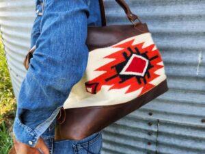 A person is standing sideways, smiling, wearing a denim outfit. They are carrying a bag with a geometric design in red, black, and white. A dog with a black and white coat is seen at the bottom right corner of the image, looking up at the person.