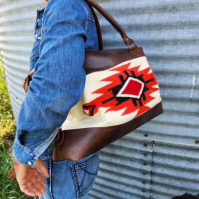 A person is standing sideways, smiling, wearing a denim outfit. They are carrying a bag with a geometric design in red, black, and white. A dog with a black and white coat is seen at the bottom right corner of the image, looking up at the person.