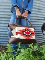 Person in denim holding a brown leather handbag with a tribal pattern. The person has visible tattoos on their hands and stands against a corrugated metal backdrop with greenery at their feet. A small part of a black and white dog is visible to the right.