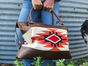 Person in denim holding a brown leather handbag with a tribal pattern. The person has visible tattoos on their hands and stands against a corrugated metal backdrop with greenery at their feet. A small part of a black and white dog is visible to the right.