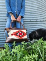 A person in a denim outfit holds a brown leather bag featuring a red, white, and black geometric pattern. They stand next to a black and white dog sniffing the green plants at their feet. The background is a corrugated metal wall.