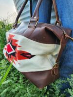 A person wearing blue jeans stands near greenery, holding a stylish bag with brown leather accents and a cream fabric middle section featuring a red, black, and white geometric pattern.