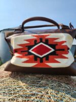 A handbag with a Native American-inspired design sits on a hay bale. The bag features a geometric pattern in red, white, and black on a beige background. The handles and corners of the bag are made of brown leather. Behind, a large round metal silo is visible.