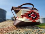 A brown leather tote bag with a colorful geometric design sits perched on a large bale of hay in an outdoor setting. The bag features red, black, and white patterns against a beige background. In the distance, blurred silos and a clear blue sky are visible.