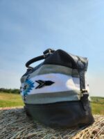 A close-up of a black and white duffel bag with a Native American-inspired geometric pattern in shades of blue and black. The bag is resting on a hay bale in a scenic outdoor setting under a clear blue sky.