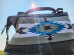 A black leather bag with an intricate, colorful woven design featuring geometric patterns in white, gray, black, and blue hues sits outdoors under the sun. Sunlight creates a lens flare effect, adding a bright spot and rays to the upper part of the image.