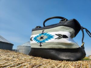 A leather handbag with a tribal patterned fabric section in blue, white, and black is placed on a hay bale. The background shows a clear blue sky and blurred silo structures.