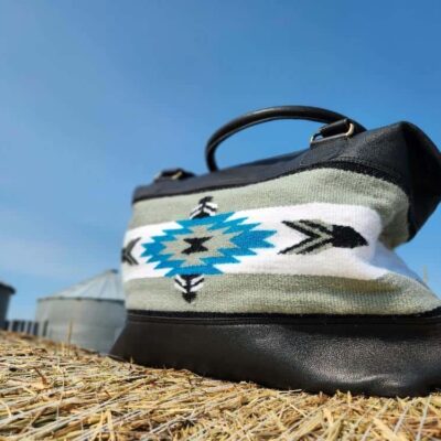 A leather handbag with a tribal patterned fabric section in blue, white, and black is placed on a hay bale. The background shows a clear blue sky and blurred silo structures.