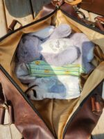 An open brown leather bag sits on a wooden surface, revealing its contents: a stuffed rabbit toy and a set of baby diapers in varying sizes. The bag has a beige interior lining and is slightly shaded.