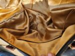 The image shows the interior of an empty handbag, lined with gold satin fabric. There are slight creases and folds in the lining, and a side pocket with a visible zipper. The bag's top is slightly opened, held by a hand.